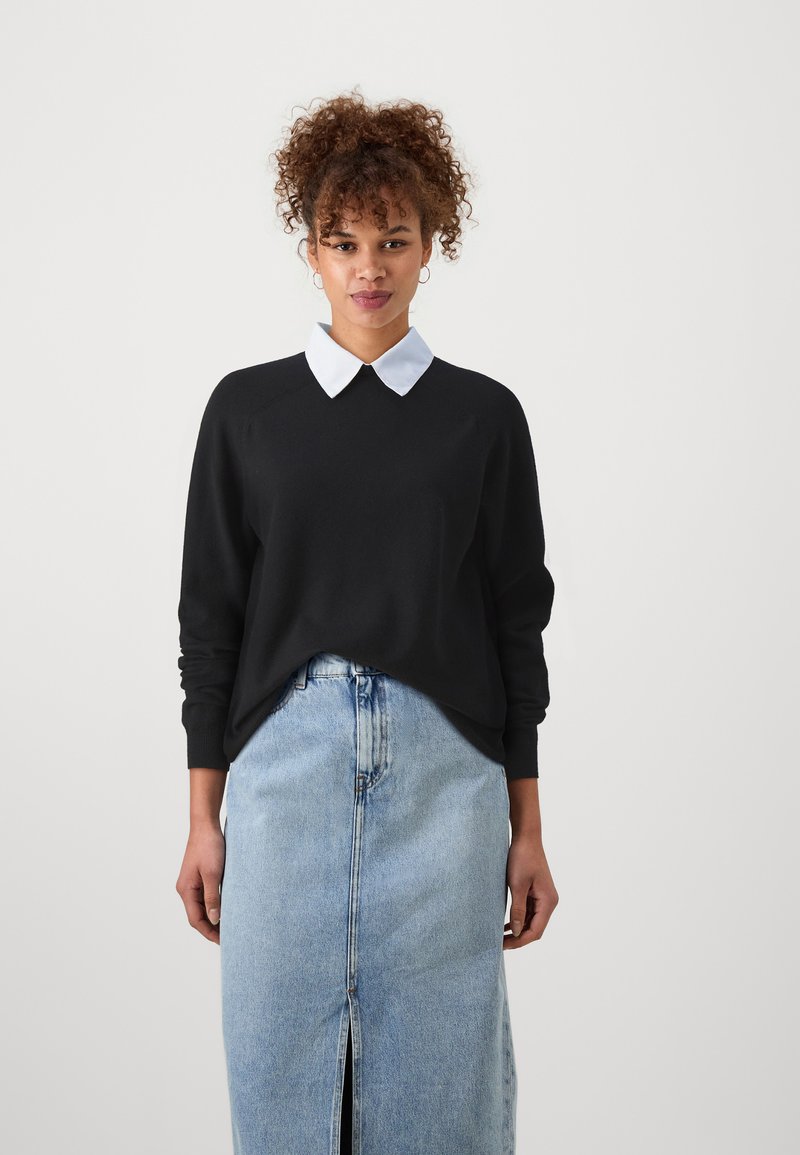 Sudadera negra con una camisa blanca de cuello debajo, con un corte relajado y mangas largas. Combinada con una falda de mezclilla azul clara.
