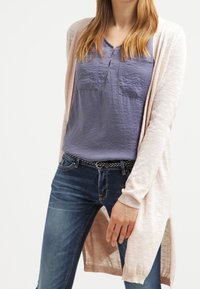 Světle růžový cardigan s dlouhými rukávy a texturovanou tkaninou. V kombinaci s volně střiženým levandulovým topem s dvěma předními kapsami. Tmavě modré džíny.