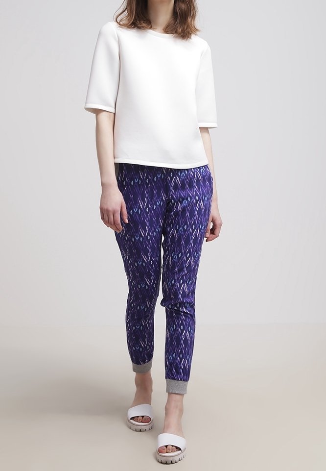 Vit kortärmad topp kombinerad med lila mönstrade byxor med diamantformer, grå manschetter och vita slip-on sandaler med en texturerad sula.