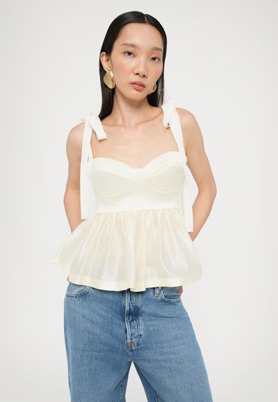 Top peplum en crema con un busto estructurado, acabado de satén suave y tirantes ajustables. Combinado con jeans de mezclilla azul claro.