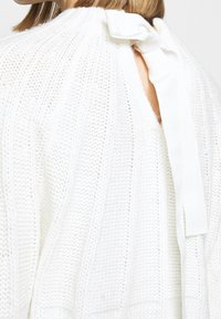 Valkoinen neulepusero korkealla kaula-aukolla, avoimella selällä ja nauha-yksityiskohdalla niskassa. Puserossa on teksturoitu kuvio, jossa on pystyribbet.