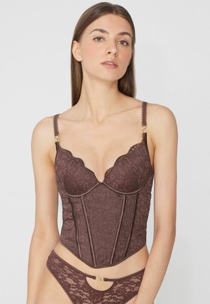 Femme portant un ensemble de lingerie en dentelle marron assorti avec un haut style corset et une culotte avec des détails anneaux dorés, posant sur fond blanc.