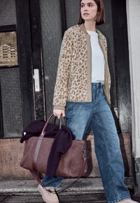 Jeune femme portant une veste imprimée léopard et un jean, transportant un grand sac en cuir marron avec un pull foncé devant un bâtiment.