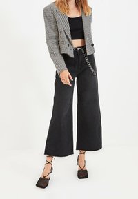 Blazer pied-de-poule noir et blanc, coupe courte, associé à un jean noir taille haute à jambes larges avec un détail de chaîne circulaire. Talons noirs à lanières.