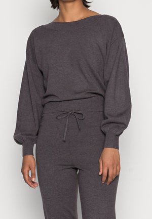 Vrouw die een donkergrijze, gebreide top met lange mouwen en ballonmouwen draagt, gecombineerd met een bijpassende high-waist broek met een trekkoord.