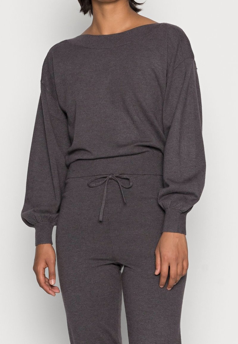 Femme portant un haut en maille gris foncé à manches longues bouffantes et un pantalon assorti taille haute avec un cordon de serrage.