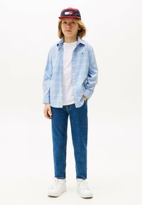 Camicia a quadri blu chiaro sopra una t-shirt bianca, abbinata a jeans di denim blu e sneakers bianche. Cappellino rosso e blu con dettaglio del logo.