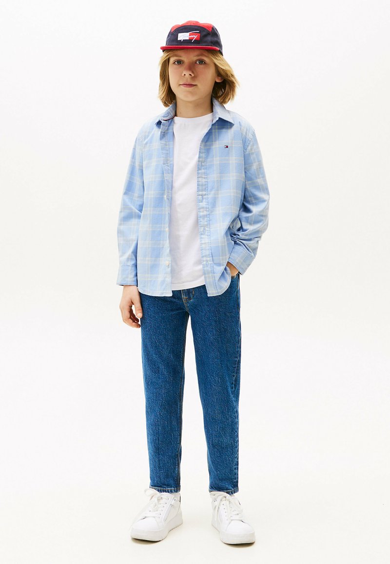 Camicia a quadri blu chiaro sopra una t-shirt bianca, abbinata a jeans di denim blu e sneakers bianche. Cappellino rosso e blu con dettaglio del logo.
