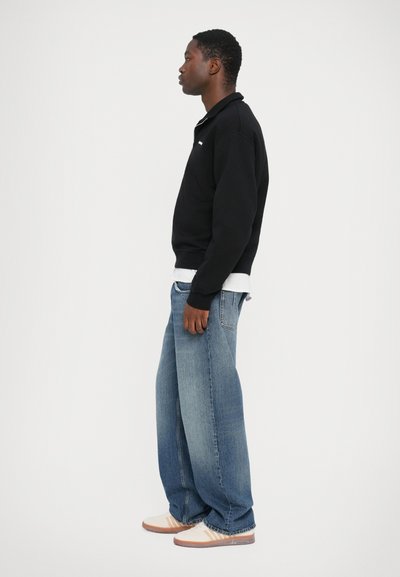 Zwarte sweatshirt, blauwe wijde jeans en crèmekleurige sneakers met bruine accenten. Eenvoudig ontwerp, zichtbare stiksels en een relaxte pasvorm.