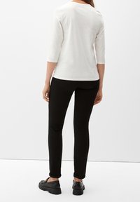 Weißes Oberteil mit drei Viertelärmeln, kombiniert mit engen schwarzen Hosen und schwarzen Schuhen. Das Outfit zeichnet sich durch ein simples Design und eine glatte Textur aus.