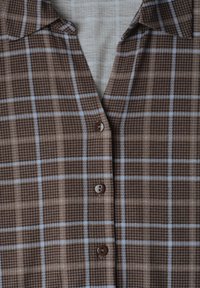 Chemise à carreaux marron et beige avec des lignes bleu clair et des boutons marron, partiellement ouverte au col.