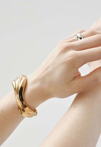 Brazalete de pulsera metálico dorado con un diseño fluido, llevado en una mano de textura suave, junto a un anillo de plata pulida en una segunda mano.
