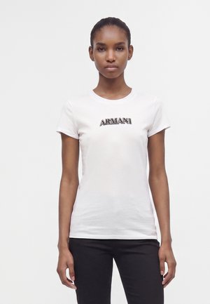 Jonge vrouw die recht vooruit kijkt, draagt een wit Armani T-shirt en zwarte broek tegen een effen witte achtergrond.