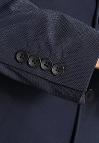 Manica di blazer texture blu navy con quattro bottoni neri. Il tessuto presenta un sottile motivo a diamante, che migliora il dettaglio visivo e la profondità.