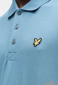 Polo azul claro feita de tecido texturizado, com um emblema de pássaro preto e dourado, pala de três botões e detalhes de gola canelada.