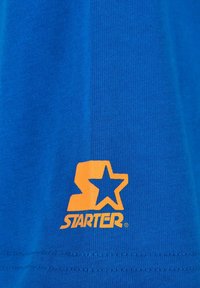 Mėlyna medvilninė marškinėlių pjesė su oranžiniu "Starter" logotipu, papildytu žvaigždės akcentu, pasižyminti lygiu paviršiumi ir raukšlėtomis kraštinėmis.