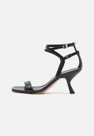 Sandale noire en cuir à talon haut avec deux brides réglables à la cheville, une bride unique à l'orteil et un talon fin et courbé.