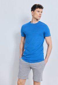 Άνδρας που φορά μπλε κοντομάνικη μπλούζα και γκρι σορτς, στέκεται με τα χέρια στις τσέπες, κοιτάζοντας προς τα πλάγια σε απλό φόντο.