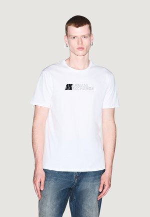 Giovane con capelli corti rossi e orecchini che indossa una t-shirt bianca Armani Exchange e jeans blu, in piedi davanti a uno sfondo chiaro e uniforme.