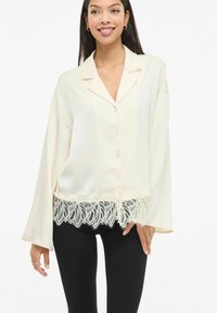 Kremfarget silkesbluse med lange ermer, en krage og intrikat svart blondedetalj langs kanten. Knappelukking midt foran.