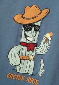 Niebiesko-szary materiał z rysunkowym kaktusem w kowbojskim kapeluszu, okularach przeciwsłonecznych, czerwonym bandaną i lodami, z napisem "CACTUS HUGS".