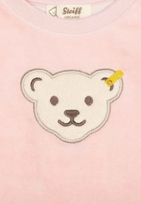 Camiseta de algodón rosa con un rostro de oso blanco bordado y acentos marrones. La oreja del oso tiene una pequeña etiqueta de tela amarilla.