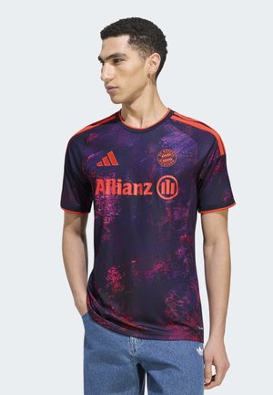 Man draagt een donkerpaarse en rode FC Bayern München voetbalshirt met Allianz-logo, staat met zijn handen in zijn zakken en kijkt naar links.