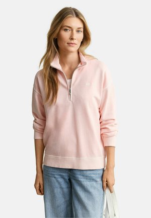 Jonge vrouw draagt een lichtroze half-ritssweatshirt en lichtblauwe spijkerbroek, houdt een witte draagtas vast met een neutrale gezichtsuitdrukking.