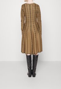 Brązowa sukienka z długim rękawem, w pionowe czarne wzory, w fasonie rozszerzanym, z długością do połowy łydki; noszona z czarnymi botkami sięgającymi kolan.