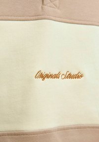 Bež i krem boje s blokovima boja, s glatkom teksturom, na kojemu je u smeđoj boji izvezena oznaka "Originals Studio". Detalji uključuju šivane šavove.