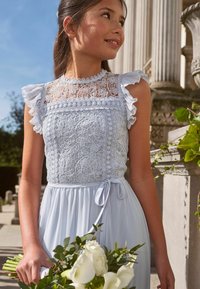Himmelblaues Kleid mit Spitzen-Bustier und Rüschensleeves, elastischem Bund und gebundenem Detail; das Modell hält einen Strauß weißer Blumen.