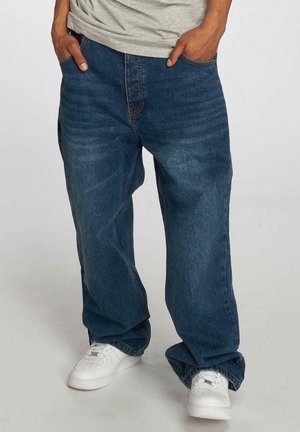 Person mit weit geschnittenen blauen Jeans und weißen Turnschuhen, die mit den Händen in den Taschen vor einem einfarbigen Hintergrund steht.