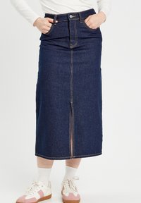 Jupe midi en denim bleu foncé avec une fente devant, cinq poches et des coutures contrastées. Associée à des chaussettes blanches et des baskets avec des accents roses.