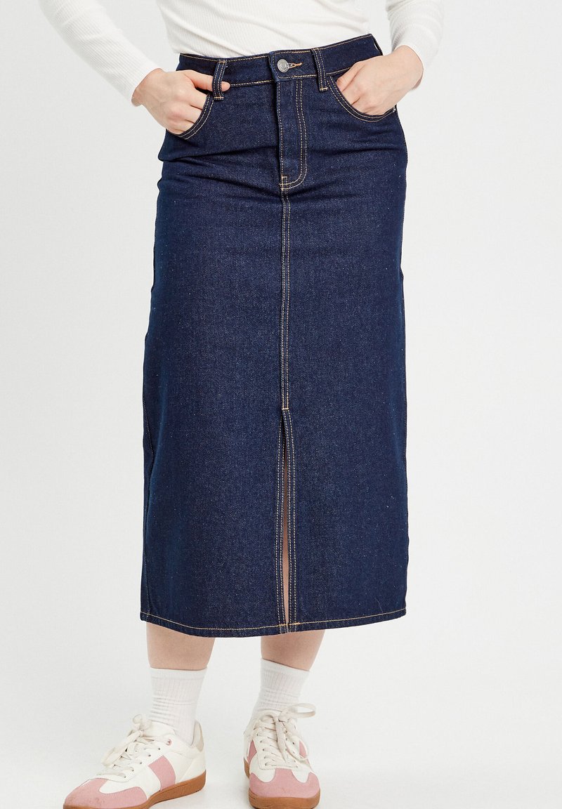 Jupe midi en denim bleu foncé avec une fente devant, cinq poches et des coutures contrastées. Associée à des chaussettes blanches et des baskets avec des accents roses.