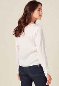 Blusa blanca de manga larga con una textura suave, escote redondo y dobladillo curvado, combinada con jeans de mezclilla oscuros con costuras detalladas.