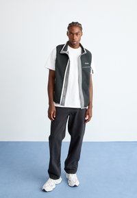 Ung mand med flettehår iført en hvid t-shirt, åben sort fleecevest, løse sorte bukser og hvide sneakers, der står på blå gulv.