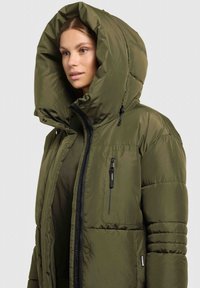 Doudoune vert olive avec une capuche oversize, une fermeture éclair sur le devant et une poche poitrine. Comprend une texture en tissu avec des sections matelassées.