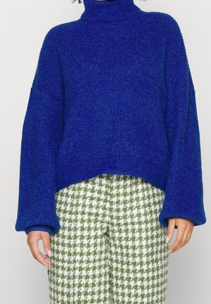 Blå stickad tröja med hög krage och vida ärmar, kombinerad med byxor i grön och vit houndstooth-mönster.
