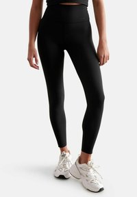 Leggings neri a vita alta realizzati in tessuto elasticizzato, con una texture liscia e un design aderente, abbinati a scarpe da ginnastica bianche.