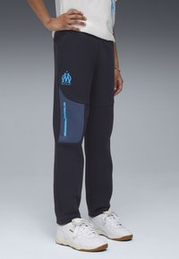 Marineblauwe joggingsbroek met een blauwe zijzak en logo. Gemaakt van zachte stof, met een elastische tailleband en taps toelopende pijpen. Witte sneakers gedragen.