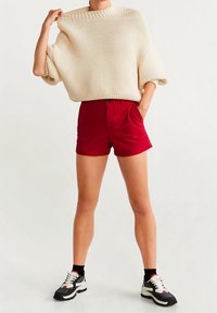 Krémový chunky pletený svetr, červené šortky a černé sportovní tenisky s bílými detaily. Pohodlný outfit s volným střihem a žebrovaným výstřihem.