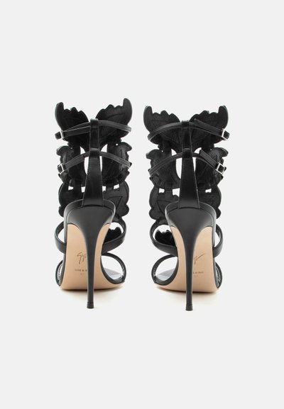 Μαύρα σανδάλια με ψηλό τακούνι, δερμάτινα λουριά σε σχήμα πεταλούδας και λεπτά τακούνια στιλέτο, εμφανιζόμενα από πίσω σε λευκό φόντο.