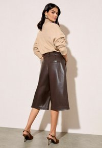 Moteris bežinėje golfiuko megztinyje, ruduose odiniuose trumpuose šortuose, rudose aukštakulnėse bateliai su atviru kulnu ir auksinėmis auskarų pareiškimais pozuoja šonu.