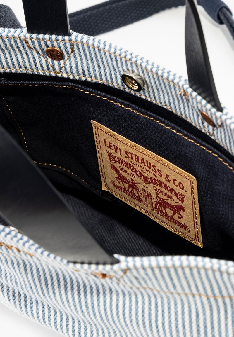 Denim- und Canvas-Shopper mit einem dunklen Innenfutter. Verfügt über ein braunes Etikett mit geprägten Grafiken und goldenen Nähten.