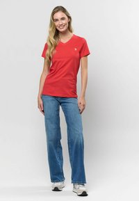 Sarkana v-veida krekls, izgatavots no kokvilnas, apvienots ar gaiši ziliem plaša piegriezuma džinsiem. Redzami ikdienišķi apavi. Uz krekla ir vienkārša logo detaļa.