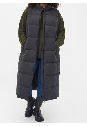 Person iført en lang sort dunvest over en grøn sweater, blå jeans og sorte støvler, stående med hænderne i vestens lommer.