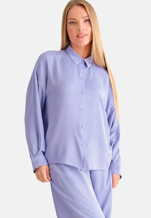 Femme aux longs cheveux blonds, portant une chemise lavande ample à boutons et un pantalon assorti, debout devant un fond clair uni.