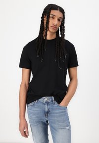 Svart t-shirt med kort ärm, rund halsringning, bärs med ljusblå slitna jeans, händerna i fickorna, avslappnad passform.