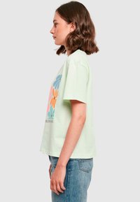 Light green cotton t-shirt with short sleeves featuring colorful flower graphics in pink, orange, blue, and green, with a round neckline.