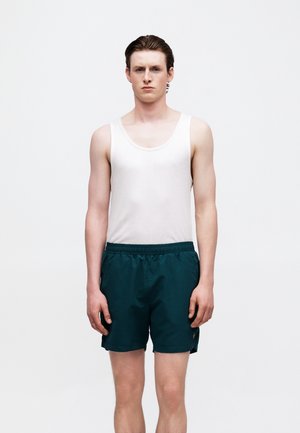 Young man with short brown hair wearing a white tank top and dark green shorts, standing against a plain white background.